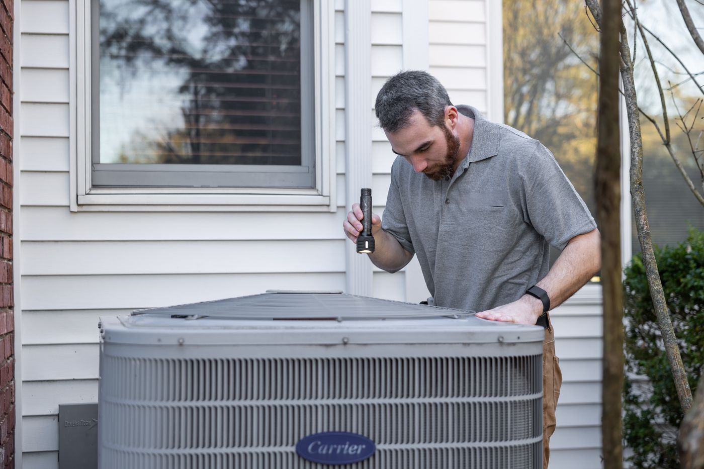 A technician with a flashlight inspecting the top of an outdoor air conditioning unit beside a house